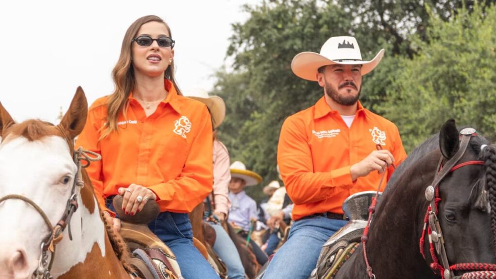 Samuel-garcia-viaja-a-disney › La Sierra Madre Two Riders in Orange Shirts on Horseback at an Outdoor Event, with a Woman Wearing Sunglasses Beside a Man in a White Cowboy Hat. › La Sierra Madre