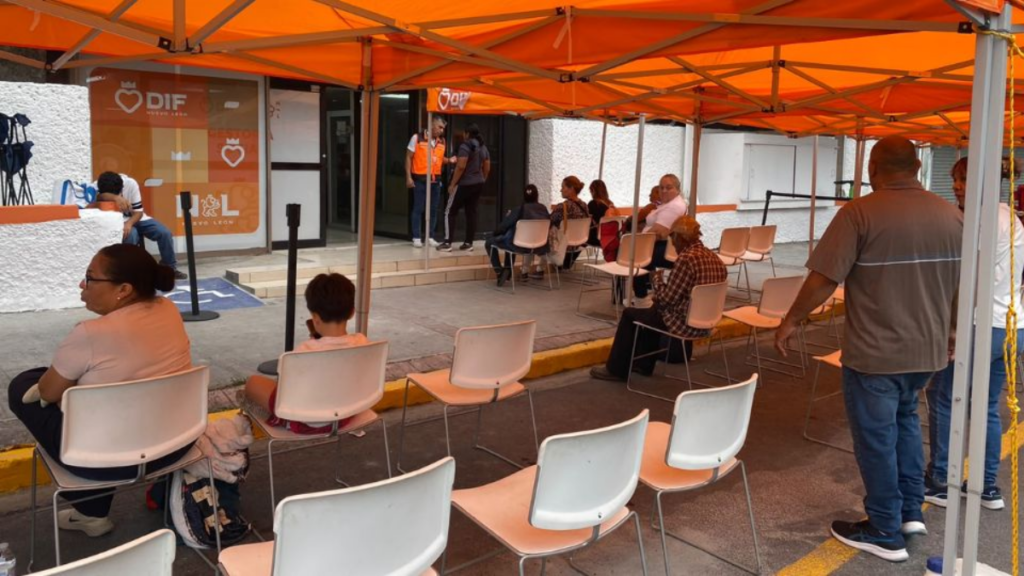 Victimas-duermen-piso-ayuda-social › La Sierra Madre People Sit in White Chairs Under an Orange Canopy Outside a Building, Waiting Near a Doorway with a Medical Organization Sign Visible in the Background. › La Sierra Madre